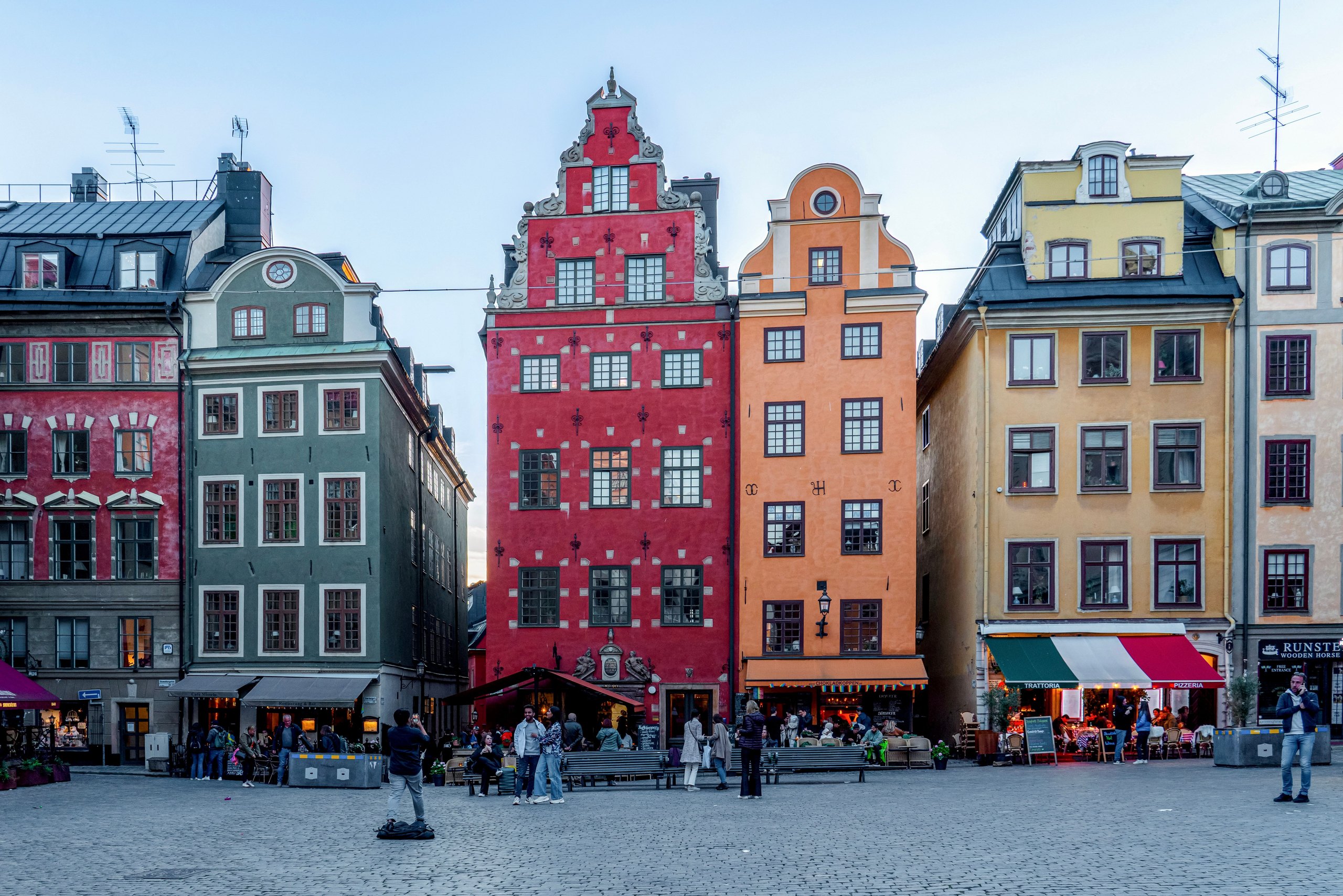 The coloured merchant houses on Stortorget in Stockholm&rsquo;s Gamla Stan &mdash; deep red, ochre and green facades with stepped gables, above cafe tables in the square.